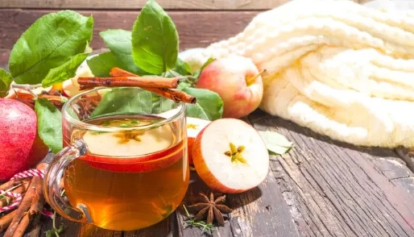 كأس من عصير التفاح المتبل مع عيدان القرفة على سطح خشبي، محاط بالتفاح المقرمش وأوراق الخريف ووشاح محبوك مريح، مما يخلق مشهد شاي الخريف المثالي.