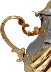 صورة مقربة لمقبض مزخرف مطلي بالذهب على إبريق شاي لامع باللونين الفضي والذهبي يتميز بتصميم معقد، ومناسب بشكل أنيق لتقديم أنواع مختلفة من الشاي.