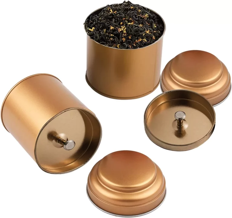تُعرض أربع علب شاي أنيقة باللون الذهبي على خلفية بيضاء نقية. إحدى العلب مفتوحة، فتظهر أوراق الشاي العطرية، بينما تظل العلب الثلاث الأخرى مغلقة بأغطيتها.