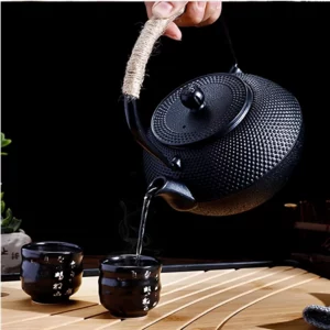 إبريق شاي أسود أنيق مع سطح محكم يسكب الشاي في كوبين أسودين صغيرين على صينية خشبية ريفية، مثالي لتجربة شاي مريحة.