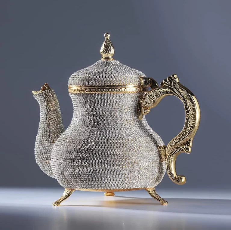 إبريق شاي مذهل مصنوع من الذهب والكريستال بتصميم معقد، يستقر على سطح عاكس أملس، مما يجعله خيارًا أنيقًا لتقديم أنواع الشاي.