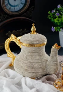 إبريق شاي فضي جميل الصنع مع لمسات ذهبية يجلس برشاقة على قطعة قماش بيضاء، مصحوبًا بساعة عتيقة ونبات في أصيص في الخلفية. مثالي للاستمتاع بأنواع مختلفة من الشاي.