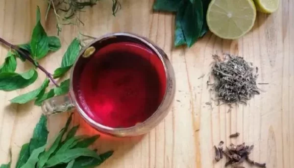 كوب من شاي الأعشاب على طاولة خشبية، محاط بأوراق النعناع الطازجة والأعشاب المجففة وشرائح الليمون والقرنفل.