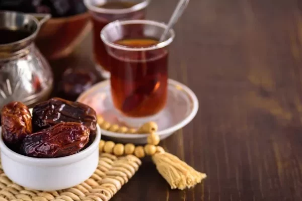 وعاء من التمر، وكأسين من الشاي التقليدي، وإبريق شاي معدني موضوع على حصيرة منسوجة فوق طاولة خشبية.