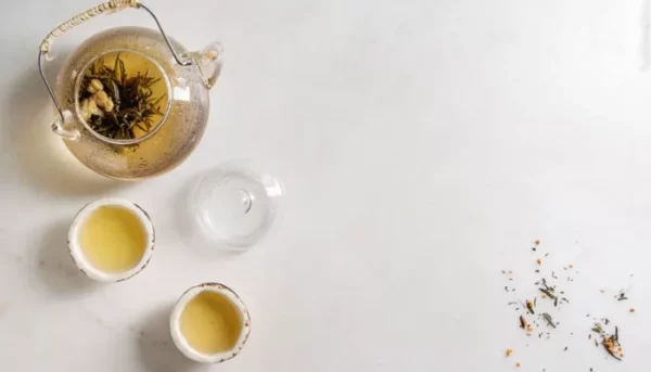 إبريق شاي زجاجي مكتوب عليه "شاي" باللغة العربية، مصحوبًا بفنجانين شاي مملوءين وأوراق شاي متناثرة على سطح أبيض.