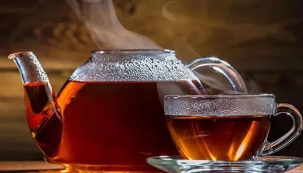 إبريق شاي زجاجي ساخن وكوب مملوء بالشاي العطري على صحن، على خلفية خشبية ريفية.