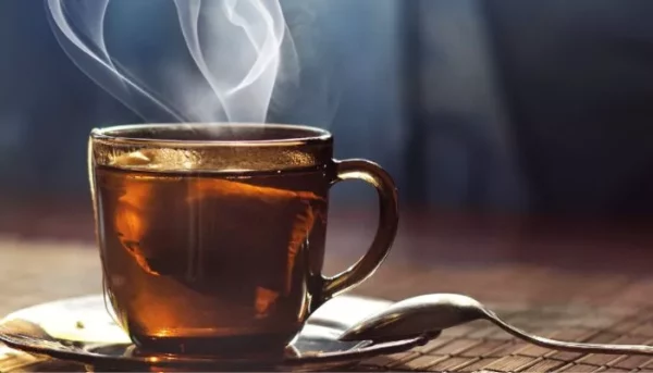 كوب زجاجي من الشاي يتصاعد منه بخار الماء، مع ملعقة موضوعة على الصحن، على سطح خشبي ريفي. مثالي لمدونة الشاي.