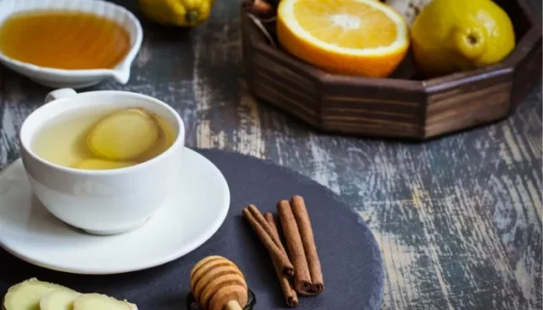 كوب شاي أعشاب أبيض مع شريحة من الليمون، محاط بالعسل وشرائح البرتقال وعيدان القرفة والليمون الكامل على طاولة خشبية.