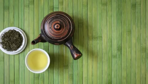 إبريق شاي سيراميكي، ووعاء به أوراق شاي سائبة، وكوب من الشاي الأخضر مرتبة على حصيرة من الخيزران الأخضر.
