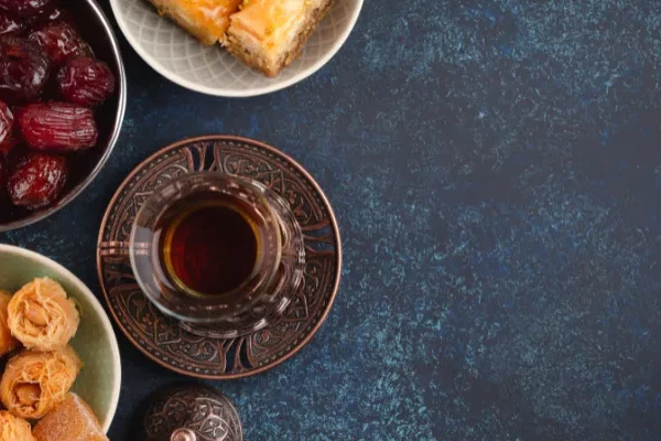 منظر من أعلى لطبق شاي نابض بالحياة يحتوي على التمر والمعجنات وكوب من الشاي العطري وحلوى متعددة الطبقات على خلفية زرقاء اللون. مثالي للاستمتاع به مع مشروبك المفضل.