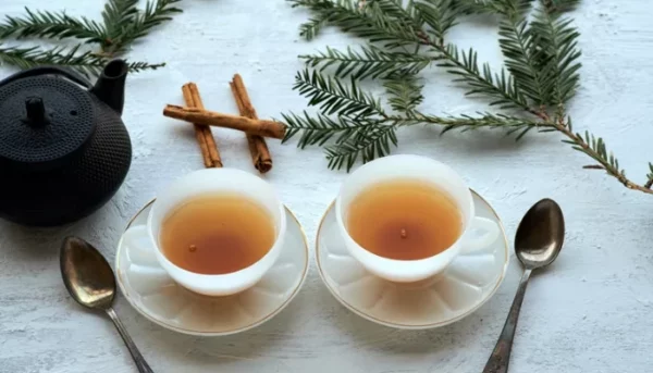 كوبين من الشاي العربي على صحون مع ملاعق، مزينين بعيدان القرفة وأغصان الصنوبر، معروضين بشكل أنيق على سطح أبيض.