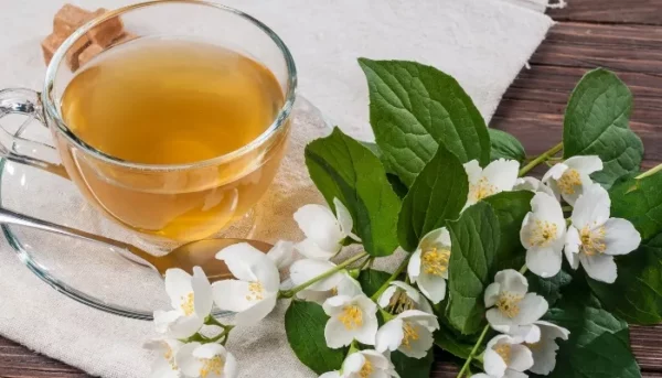 كوب من الشاي العطري على صحن مع ملعقة، مصحوبًا بأزهار الياسمين الطازجة وأوراق خضراء نابضة بالحياة على طاولة خشبية ريفية.