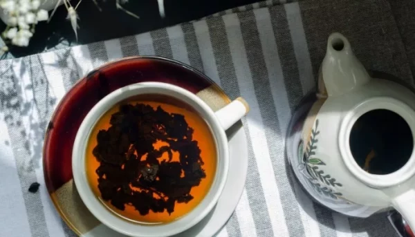 كوب من الشاي بأوراق عائمة بجانب إبريق شاي مزخرف بشكل معقد على قطعة قماش مخططة ملونة.